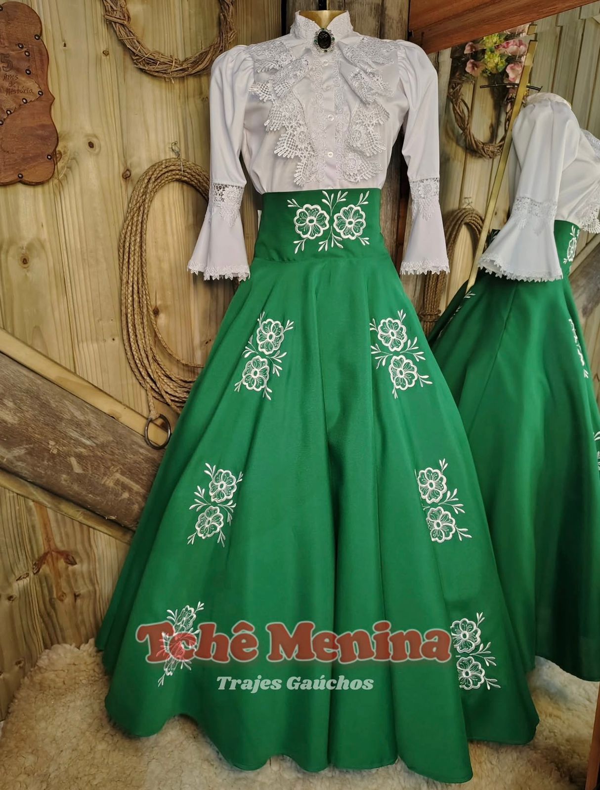 Prenda com vestido rosa e marinho bordado num cenário de CTG com roda de carro e bandeira do Rio Grande do Sul